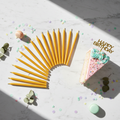 24 slim golden beeswax candles fanned out on marble surface next to cake slice and confetti