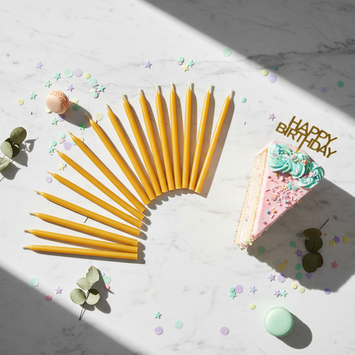 24 slim golden beeswax candles fanned out on marble surface next to cake slice and confetti